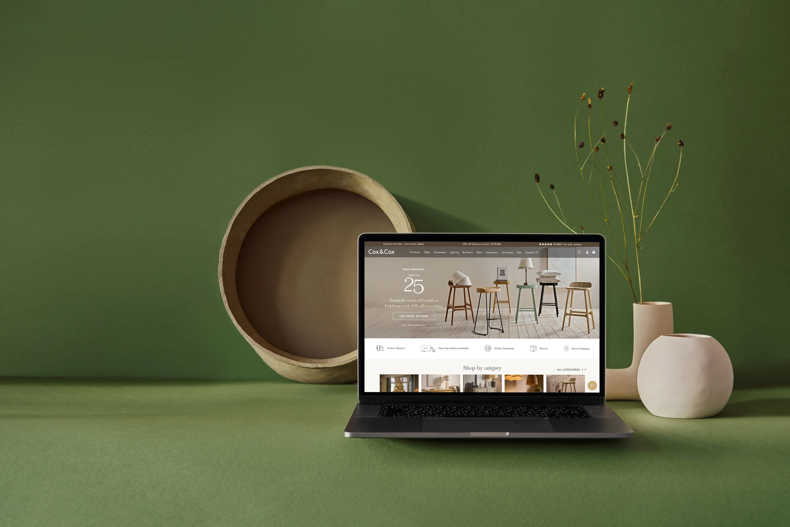 Cox & Cox interiors store displayed on a MacBook beside ceramic vessels against a green wall.